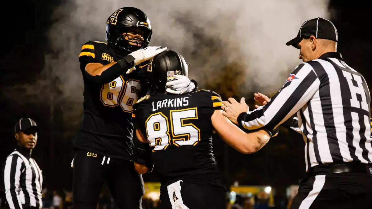 Appalachian State football players Kanen Hamlett and David Larkens celebrate, flanked by referees, with smoke swirling in the background.