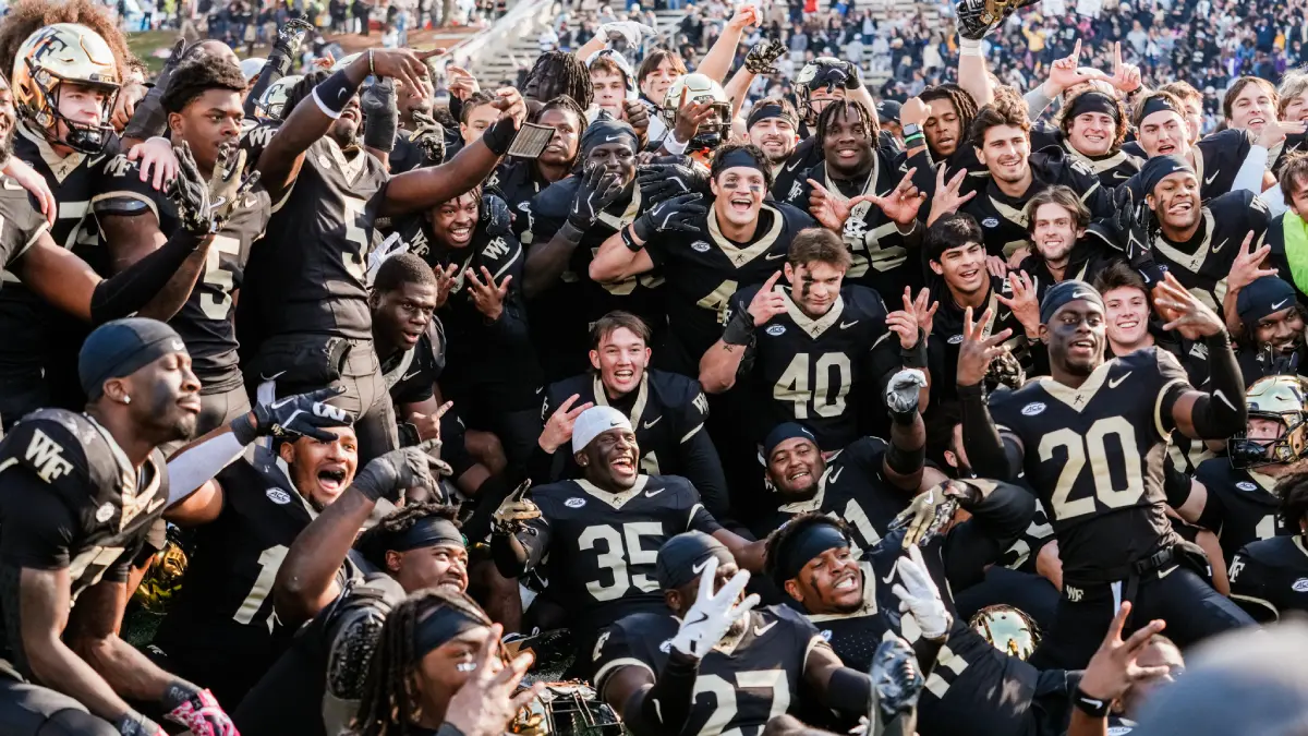 College football team in black and gold uniforms celebrating together on the field, arms raised in triumph.
