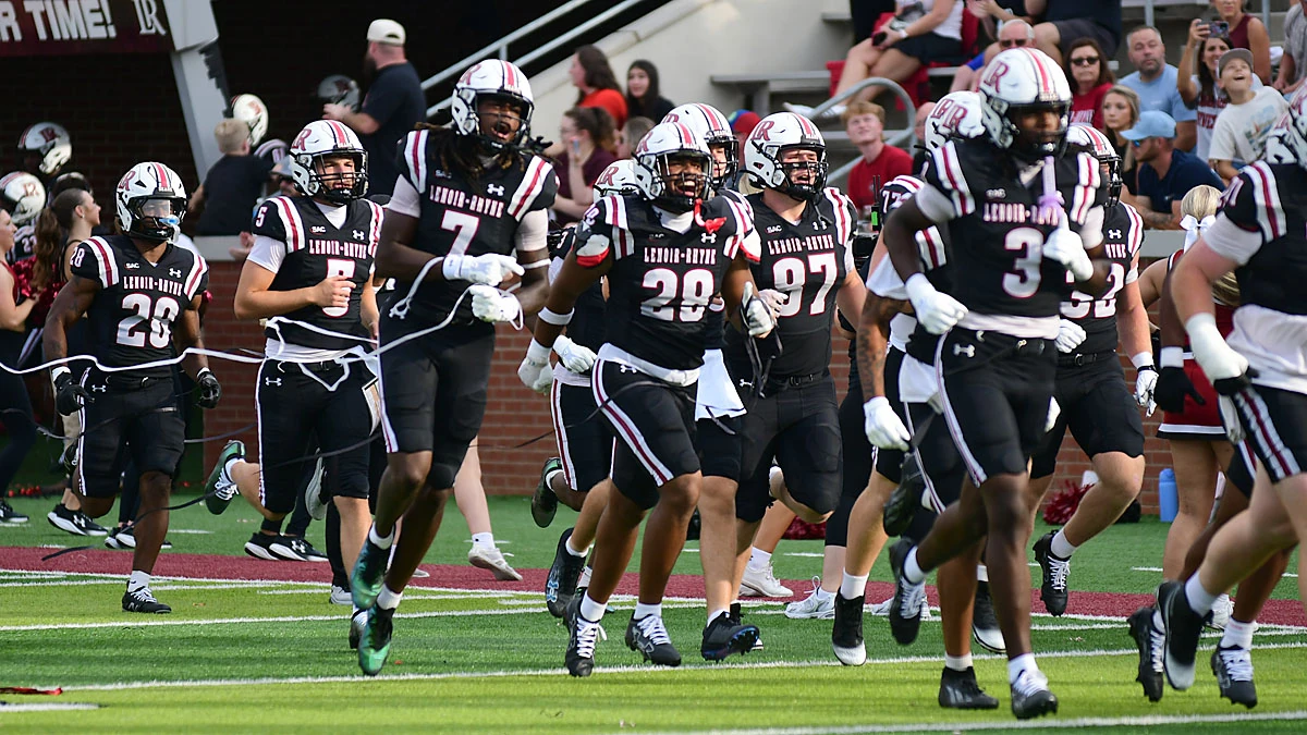 Lenoir-Rhyne football players run onto the field in black uniforms as crowds cheer in the stands