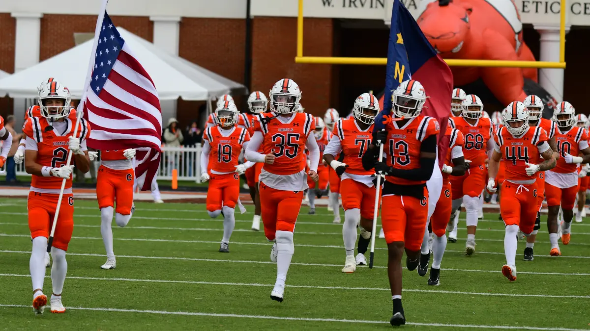Campbell University football players charge onto the field before a home game carrying US and North Carolina state flags.